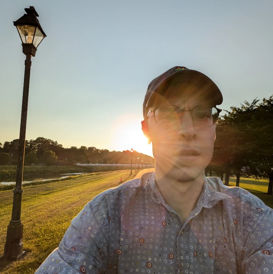 a photograph of Brandon Fannin squinting into the sun, wearing a ball cap, on a bike path, surrounded by trees